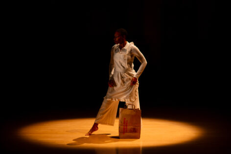 Marjani in white shirt, pants and apron walking away from a brown paper grocery bag. One heel is lifted as her hands cling to her clothes. A moment from Blondell Cummings' Chicken Soup @ Danspace Project. Photo by Rachel Keane.