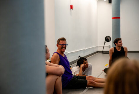 Neil, a white man with short brown hair, is sitting on the floor and smiling goofily at something he seeing beyond the camera frame. He wears a purple tank top and black shorts. Four workshop participants are sitting on the floor surrounding Neil. One participant looks at Neil, while two others are looking off camera and are smiling. The back of the fourth participant's head is pictured, nearest to the camera. Photo by Rachel Keane.