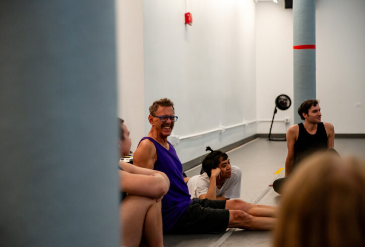 Neil, a white man with short brown hair, is sitting on the floor and smiling goofily at something he seeing beyond the camera frame. He wears a purple tank top and black shorts. Four workshop participants are sitting on the floor surrounding Neil. One participant looks at Neil, while two others are looking off camera and are smiling. The back of the fourth participant's head is pictured, nearest to the camera. Photo by Rachel Keane.