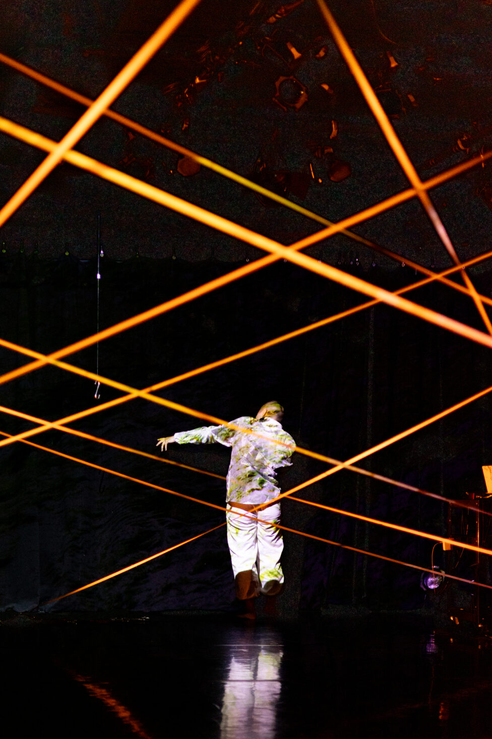 ID: Sha stands in the middle of dark stage in a white co-ord with green paint splotches on it. Orange wires cross over each other on stage. Photo: Kade Joy