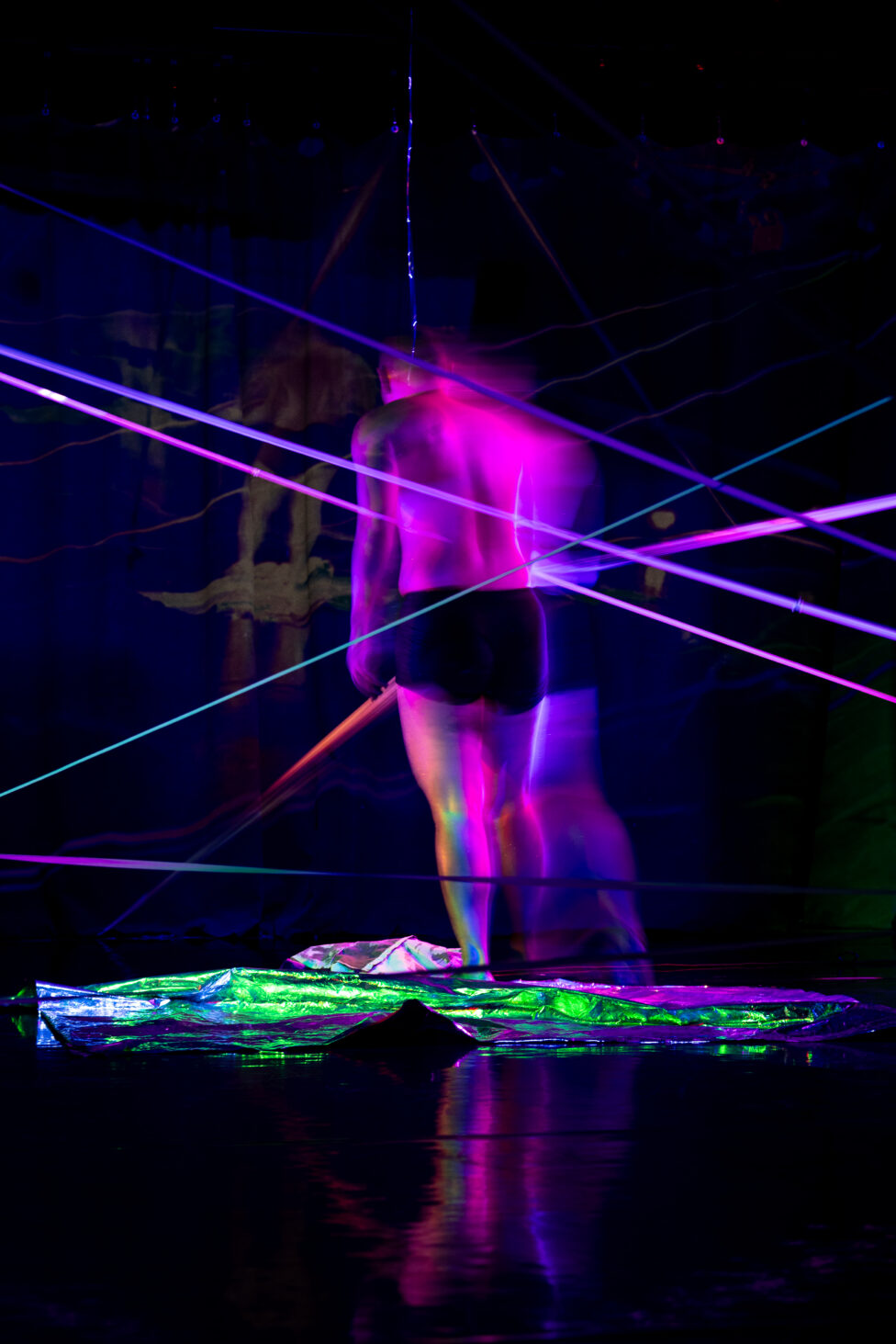 ID: Sha stands in the middle of a dark stage with tight black shorts on. Purple, pink, and blue wires cross the stage, and green-lit aluminum is on the stage floor. Photo: Kade Joy