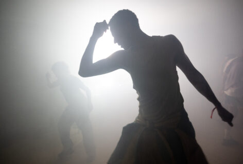 A silhouetted figure moves dynamically in a hazy, backlit space, their arm raised toward their head and body angled forward with intensity. The air appears thick with fog or humidity, diffusing the bright light behind them. Two additional figures are visible in the background, partially obscured, also engaged in movement. The image conveys exertion, atmosphere, and collective physical practice emerging through heat, density, and motion. Photo by Nir Areli.