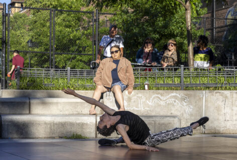 Dancer wearing black shirt, black and white pants, sitting on the ground and leaning back looking to the sky. In performance in Travers Park, Queens. Joseph Pinlac- Queensboro Dance Festival 2024