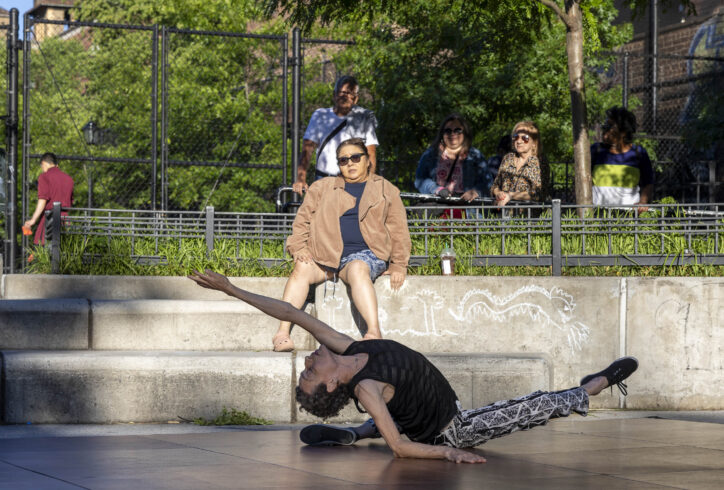 Dancer wearing black shirt, black and white pants, sitting on the ground and leaning back looking to the sky. In performance in Travers Park, Queens. Joseph Pinlac- Queensboro Dance Festival 2024