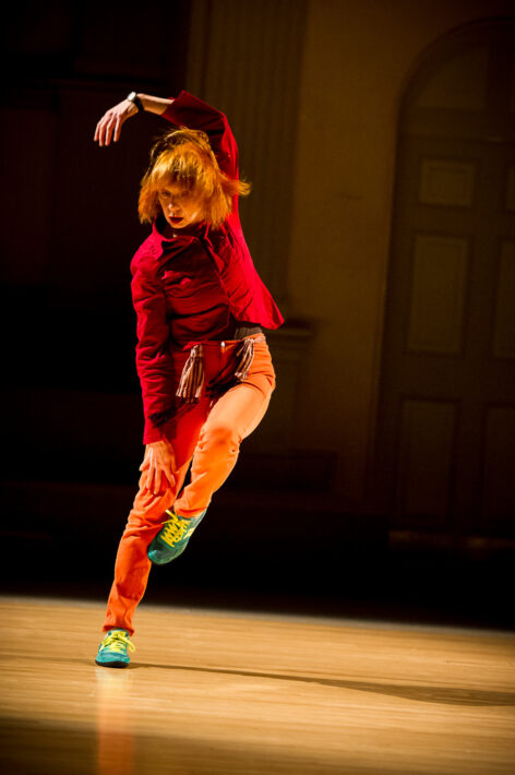 Woman in performance wearing red shirt, orange jeans and blue shoes with tassels around waist, falling slightly sideways. Photo by Ian Douglas.