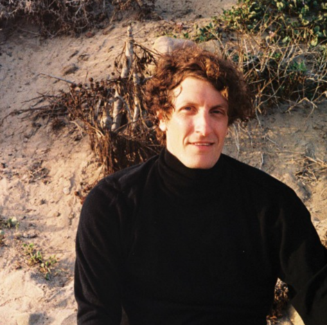 A man with curly hair sits outdoors against a sandy, natural backdrop, wearing a black turtleneck. He faces the camera with a calm, slightly introspective expression in warm, natural light.