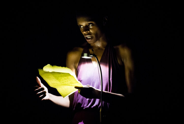 Okwui Okpokwasili reads from a brightly lit paper in a darkened room. She is a Nigerian woman wearing a purple top. Photo by Ian Douglas.