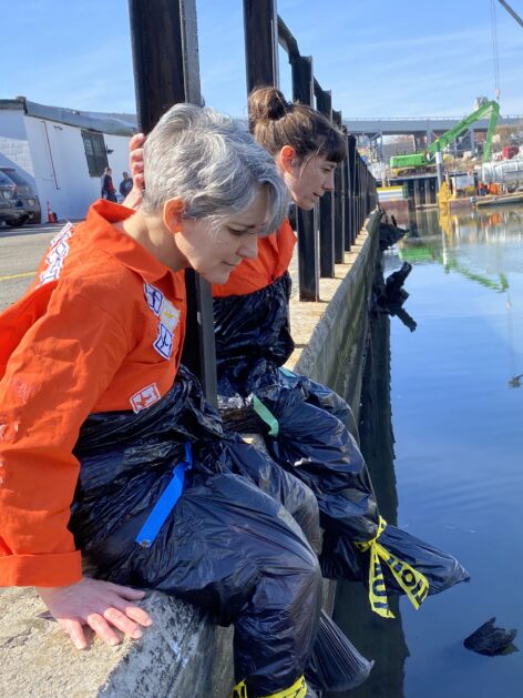 Clarinda (in front) (a person with short salt and pepper hair and pink skin) and Carolyn (a white woman with dark brown hair in a ponytail), in profile, sit on the edge of a concrete barrier, beneath a short black fence. They are wearing bright orange long sleeves on top and pants made of garbage bags held together with caution tape below the waist. Below them is the Gowanus Canal, calm and reflecting the blue sky, and they are looking into it. Small, in the background, is the BQE and a bright green crane. Photo by Kelly AuCoin.