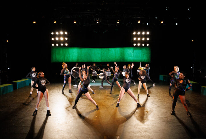 2 majorette dance teams dressed in black, blue, and green leotards and jumpsuits dance in front of a marching band on a large theatrical stage. There is a hanging green turf covered wall behind them, as well as football stadium lights. Photo by Maria Baranova