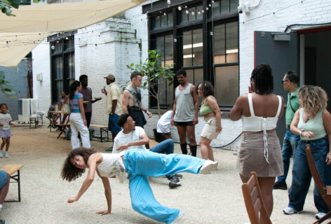 People gathered in open courtyard. Dancer is in motion at a low level with a leg and an arm in the air. Aisha Bee Photography.