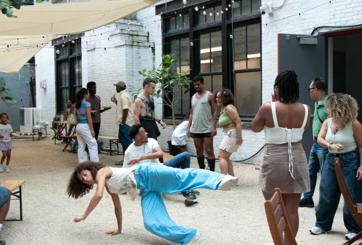People gathered in open courtyard. Dancer is in motion at a low level with a leg and an arm in the air. Aisha Bee Photography.