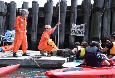 Clarinda and Carolyn, wearing orange jumpsuits, are on a raft near the wooden pylons of a pier in the middle of a body of water (Flushing Creek). Clarinda is standing to the left of the image, hands in pockets. Carolyn, wearing a big yellow elbow-length glove, is squatting to the right, showing a group of people in colorful kayaks something from a cage that is attached to the raft. Both artists are wearing yellow caution tape at their ankles.