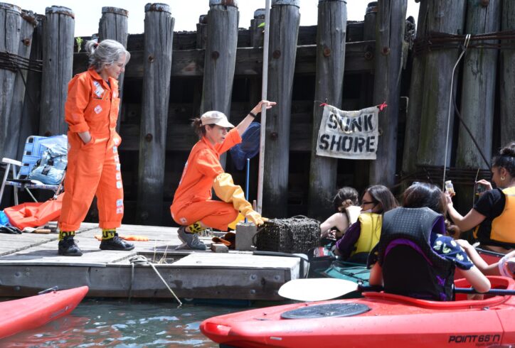 Clarinda and Carolyn, wearing orange jumpsuits, are on a raft near the wooden pylons of a pier in the middle of a body of water (Flushing Creek). Clarinda is standing to the left of the image, hands in pockets. Carolyn, wearing a big yellow elbow-length glove, is squatting to the right, showing a group of people in colorful kayaks something from a cage that is attached to the raft. Both artists are wearing yellow caution tape at their ankles.