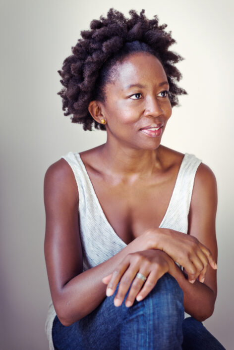 Wanjiru, a Black woman with short hair, sits with her hands on her left knee. She wears a grey tank top and jeans. photo © Mathieu Waddelle