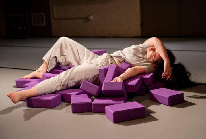 Sofia, a white femme wearing grey swaeats, lies on a pile of purple yoga blocks. Photo by Rachel Keane.