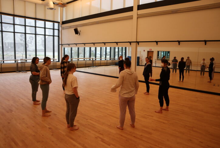 Chloë Nagle Çetinkaya teaches undergraduate dancers at the University of Wisconsin-Stevens Point. Everyone is gathered in a circle. She is on the right-hand side wearing all black with a white KN95 mask. Photo by Kevin Obsatz.