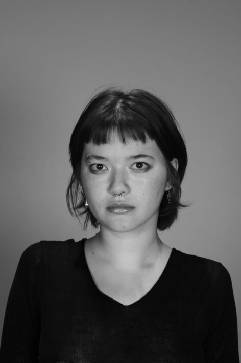 Photo of young woman with short hair and bangs in front of a plain background, wearing a black knit sweater. Photo credit: Moksha Akil.