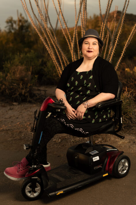 Naomi Ortiz, a light-skinned Mestize, sits in their scooter in front of an ocotillo in the desert. They are wearing a fedora hat, hoop earrings, black dress with cacti print and pink boots. Photo Credit: Jade Beall