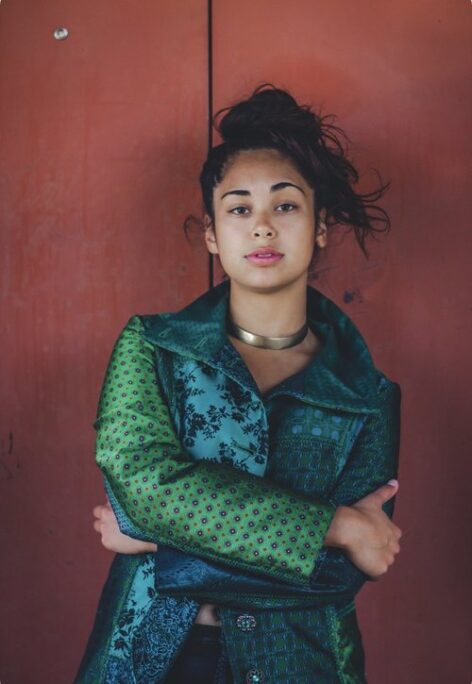 A photo of AJ Castro. A medium-skinned person poses with their arms crossed in front of a rusty-red background. They’re looking straight at the viewer with a raised eyebrow. They have long dark hair in a windswept bun on their head. They’re wearing a dark-green coat made of patchwork pieces of brocade fabrics. Photo courtesy of the artist.