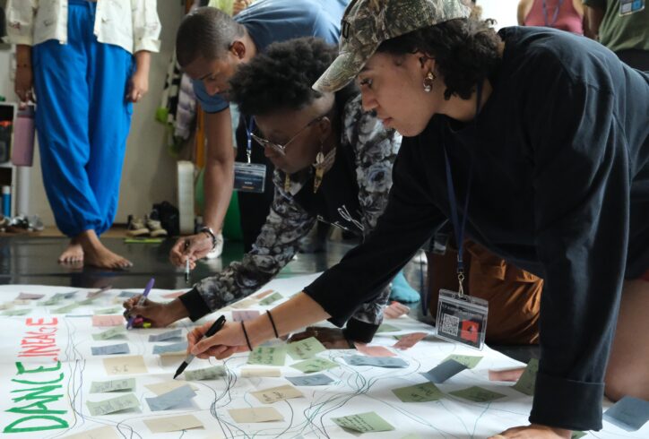 Black dancers at the BDCM Summer Untensive lean over a large floor poster, writing notes and adding sticky notes as they map their Black dance lineage together in community. Photo by Martin Nuñez-Bonilla.