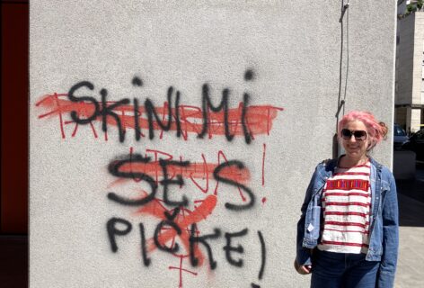 luciana standing by a graffiti in Ljubljana, Slovenia, She has wide legged jeans and birkenstock sandals a Mexican Chiapas embroidered shirt with an open jean jacket on top. She has a big smile dark sunglasses and pink hair pulled back. The graffit is in Slovenian: