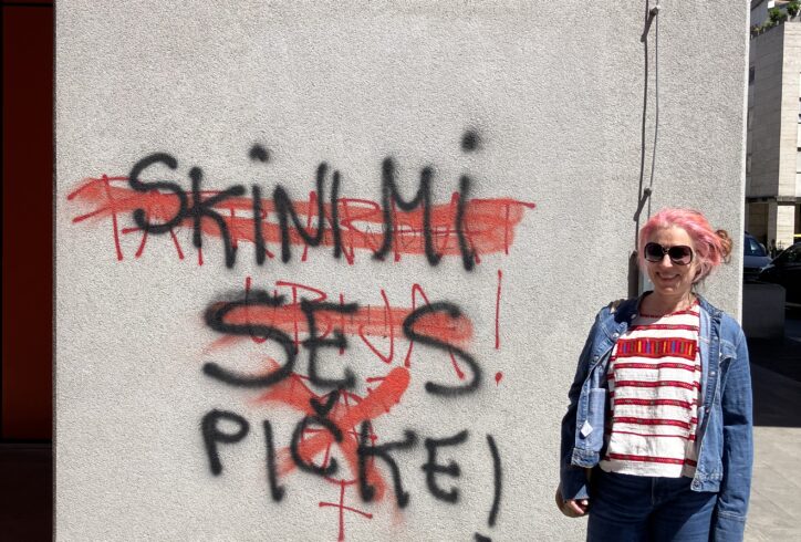 luciana standing by a graffiti in Ljubljana, Slovenia, She has wide legged jeans and birkenstock sandals a Mexican Chiapas embroidered shirt with an open jean jacket on top. She has a big smile dark sunglasses and pink hair pulled back. The graffit is in Slovenian: 