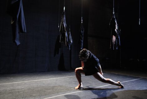 A dancer in a black outfit performs a low lunge on a dark stage with several garments hanging suspended in the background. Photo by Theo Cote.