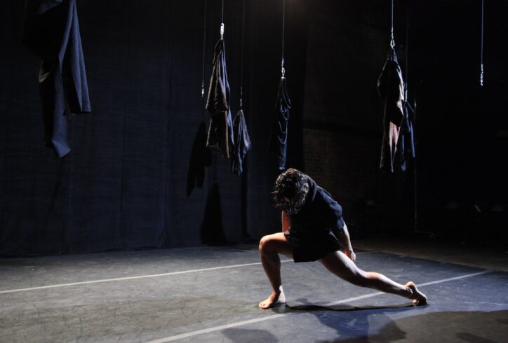 A dancer in a black outfit performs a low lunge on a dark stage with several garments hanging suspended in the background. Photo by Theo Cote.