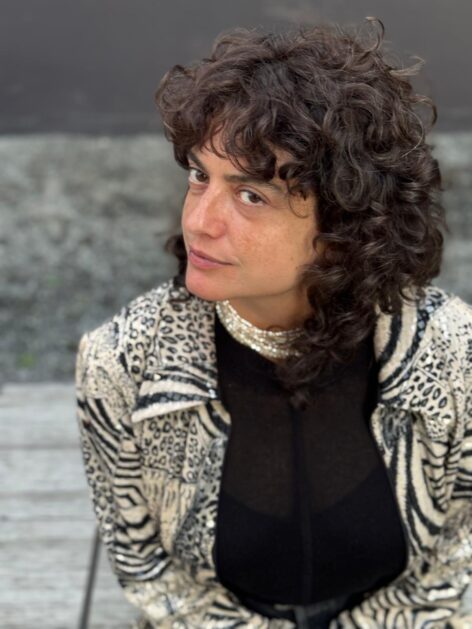 The image shows Maka, a white Caucasian woman with mid-length curly hair. She is wearing a jacket decorated with abstract beige and black patterns over a black shirt, paired with a pearl necklace worn close to the neck. Her face is captured in a three-quarter profile, and she is looking upward toward the camera. Photo by Christian Rosche.