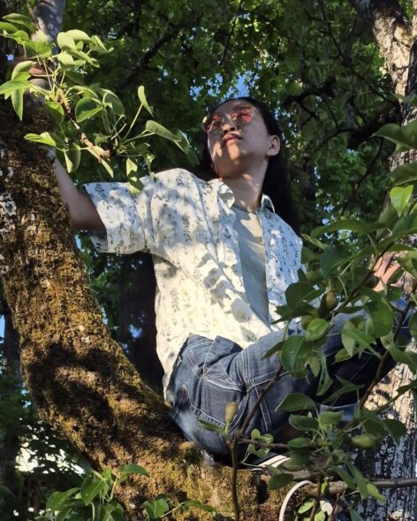 A photo of Nic Chang. A medium-skinned person sits in a pear tree gazing upward to the left into the branches. He has long black hair and reflective red-tinted glasses. He’s wearing a white short-sleeve shirt with a botanical pattern unbuttoned over a light green striped shirt and jeans. He is reaching up into the branches.