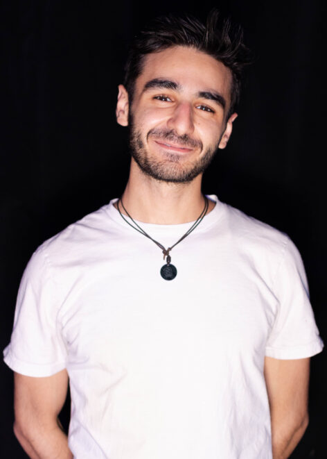 A photo of Moqu Alqudah. He grins at the camera and wears a white t-shirt and a black necklace. Photo courtesy of the artist.