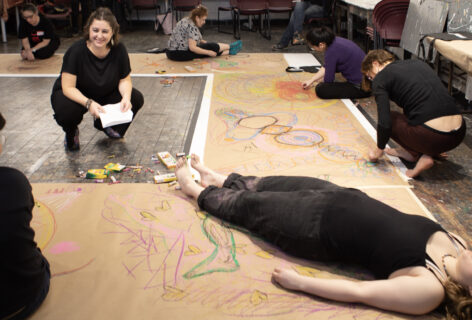 A community art workshop in progress, with participants gathered on a studio floor covered in large sheets of paper. In the foreground, a person lies on a body-sized drawing while colorful outlines, symbols, and abstract marks are created around them with crayons and pastels. Several participants kneel or sit nearby, drawing and collaborating on large-scale body maps. At the center, workshop leader Anabella Lenzu crouches with a smile, holding papers and engaging with the group. The room contains chairs, tables, and art materials, creating an atmosphere of creativity, reflection, collaboration, and embodied artistic exploration. Photo by Todd Carroll.