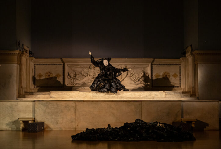 A photo from Maria Duarte Quiroga's performance for Movement Research at the Judson Church. A dancer stands at the altar dresses in all black with a white mask on their face. Photo by Rachel Keane.