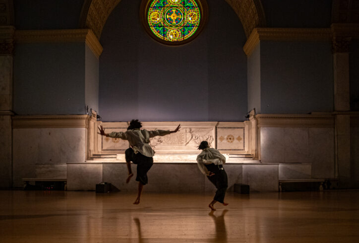 Photo from Stephanie Hewett's performance from Movement Research at the Judson Church. Two dancers move in the space. One dancer steps forward while the other is caught mid leap. Photo by Rachel Keane.