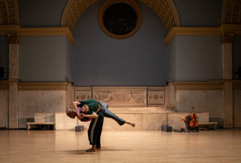 Paul Singh performs for Movement Research at the Judson Church. Paul supports a dancer as they lay over Paul's back. Paul stands in a deep squat. Photo by Rachel Keane.