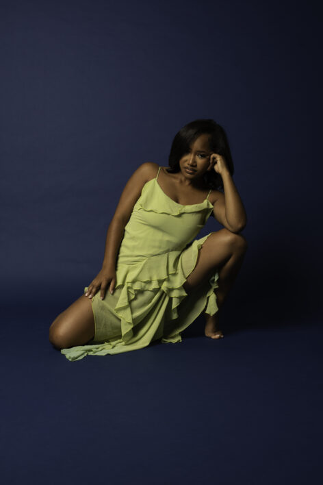 Rochelle Phillips poses in a studio against a deep navy blue backdrop, wearing a flowing chartreuse-green dress with delicate spaghetti straps and layered ruffles. She sits low to the ground in a sculptural pose, with one knee bent and the other raised, resting her head on one hand while looking directly at the camera. Her shoulder-length dark hair is styled in a sleek side part. Soft studio lighting highlights the texture of the dress and the contours of her pose, creating a refined, contemporary portrait with a calm, confident, and introspective mood. Photo by Ta Nycia Wooden.