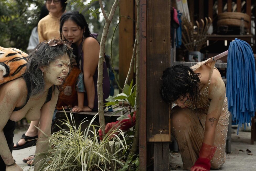 Jas Lin's Performance at 柚園自然生活節 (Pomelo Garden Natural Living Festival) in Pingtung, Taiwan with 黃藍 Huang Lan. Two performers positioned on either side of a wooden wall, both knelt on the floor, one looks down, one snarling at the wall. They are surrounded by and clad in a chaos of mixed materials, natural and man-made. Photo by Lingbin Zhong.