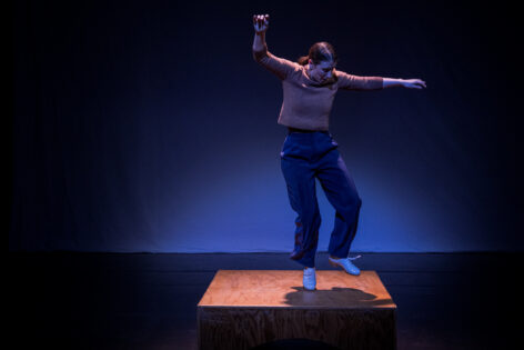 A dancer with light blue tap shoes on in mid air with their arms outstretched dancing upon a small platform. Photo by domenik lee.
