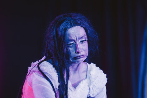 A close-up portrait of Andrea with a long dark wig and heavy theatrical makeup under dramatic blue and magenta stage lighting. Their face is scrunched into an exaggerated pout, with furrowed brows and downturned lips, conveying sadness, disappointment, and melodrama. Photo by Sanchez Productions.