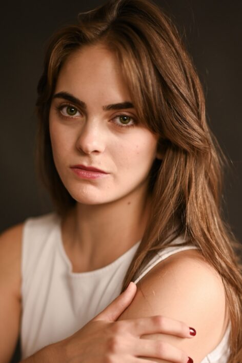 Young woman in white shirt with long brown hair looking into camera with hand on shoulder. Photo by Milan Furtado.