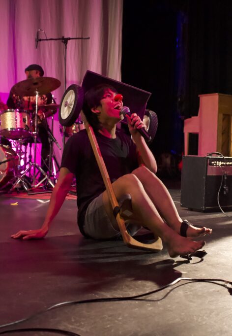 Image of an young male-presenting Asian person on stage. They are wearing a red clown nose and holding a mic. They are sitting on the stage. They are trapped inside an upside down harp cart. Photo by Tim Nohe.