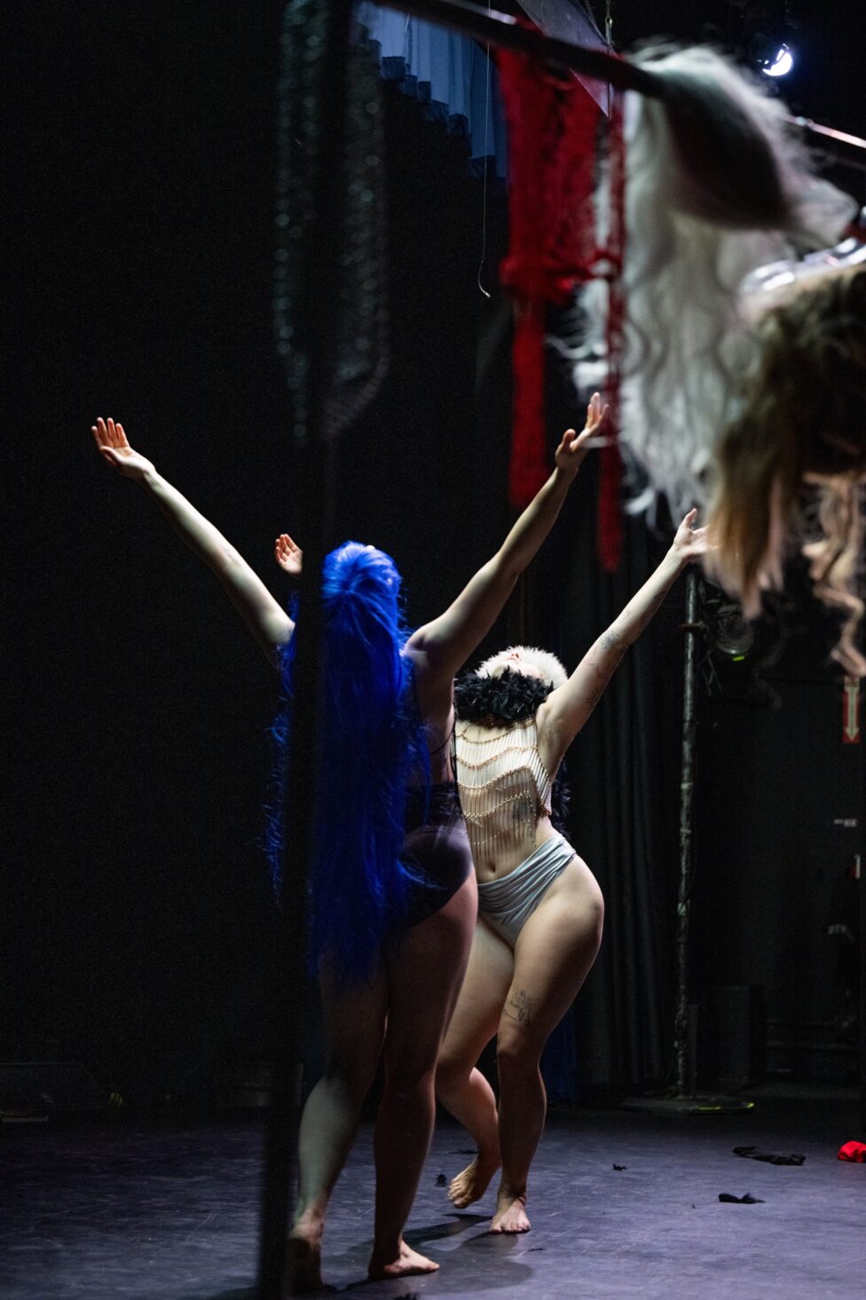 “Wanting Practice,” choreographers Lena Martin and Mak Thornquest. Photographed from behind a clothing rack, two bodies step towards one another, one in a long blue wig and one in a fur hat and black feather boa, both with arms extended upwards in a Y position. Photo by Josh Rose courtesy of Ghostlight Residency.