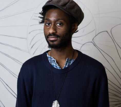 A portrait image of Donovan taken by Olivia Wein. He’s wearing a blue and white checkered long sleeve shirt underneath a navy blue sweater. Here he is also wearing a brown plaid newsboy cap and a silver necklace. Photo by Olivia Wein.