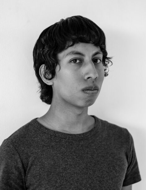 A black and white portrait of the artist. He wears a dark crewneck tee and stares directly into the camera. Photo by Santiago Sánchez.