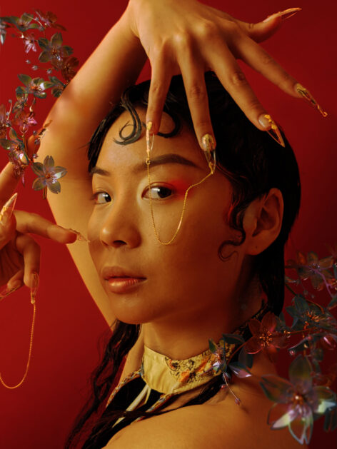 Jas Lin poses in front of a deep red backdrop. Their hands gesture around their face with gold-ornamented nails. Green and purple flowers decorate the top left and bottom right corners of the image. Photo by Andrew Thomas Huang.