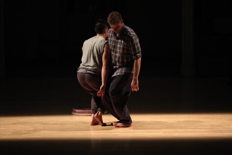 Within a stage lit theatrical setting, two figures stand close together facing in opposite directions with their shoulders touching and their gaze looking downward. Photo by Paula Court.