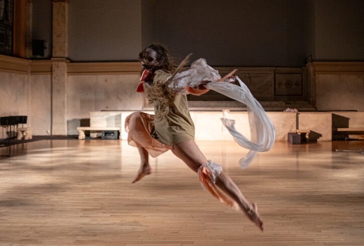 Photo from Ellen Söderhult's performance for Movement Research at the Judson Church. A dancer leaps through the air. They have face paint on and trail fabric behind them in the air. Photo by Rachel Keane.