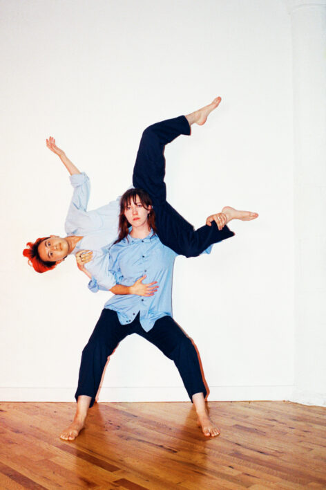 Miranda and Noa wear light blue button up shirts and navy blue pants against a white wall and wood floors. Noa rests sideways on Miranda's shoulders. Photo by Carson Stachura.