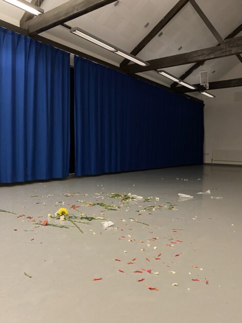 A view of a dance studio with electric blue curtain, brown beams and a light grey dance floor. The floor is covered with a scattering of flower petals in red and yellow, leaves and tissue paper. Photo courtesy the artist.