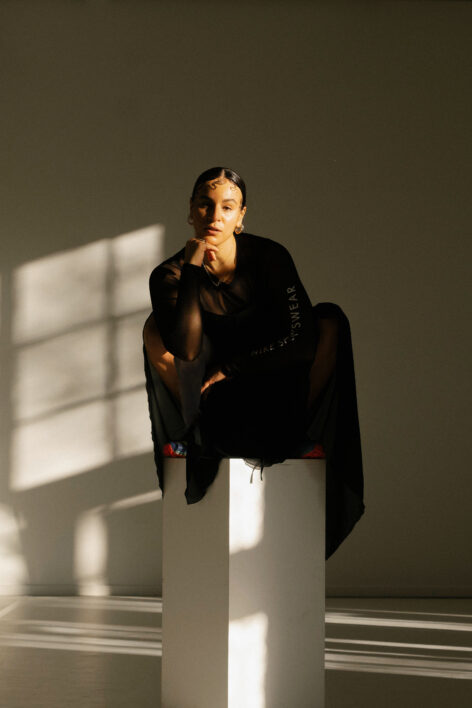 Dancer sitting on a white high podium in a dance studio with white walls - dramatic shadows in the room. Photo by Senay Berhe.